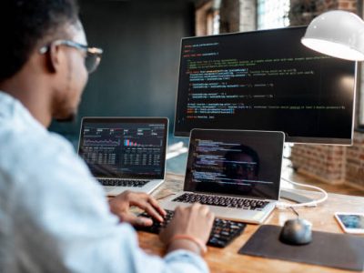 Young african male programmer writing program code sitting at the workplace with three monitors in the office. Image focused on the screen