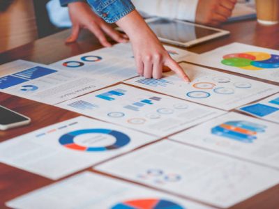 Paperwork and hands on a board room table at a business presentation or seminar. The documents have financial or marketing figures, graphs and charts on them. There is a digital tablet and laptop on the table