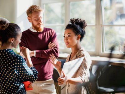 Colleagues standing in a small group discussing something. One of the women is holding documents and gesturing with her hands as the others watch and listen.